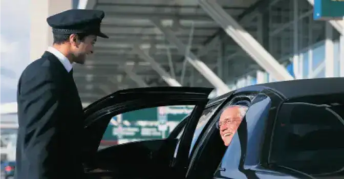 A smiling chauffeur in a black uniform and cap opens the door of a sleek black sedan for a passenger inside a luxury vehicle at an airport terminal.