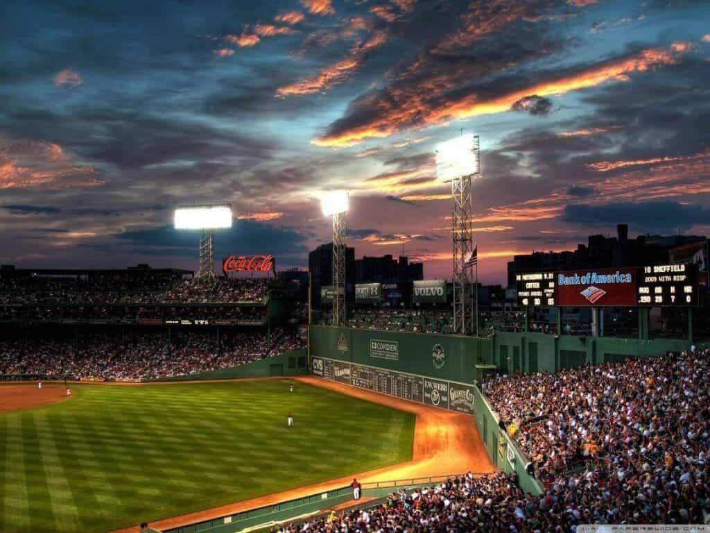 Fenway Park baseball stadium at sunset with bright floodlights, a lush green field, and a packed crowd in Boston, Massachusetts
