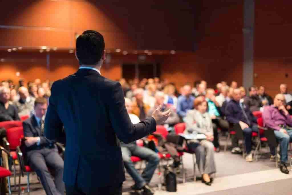 A man wearing a dark suit addresses a seated audience in a conference hall; rows of red chairs and attendees in business attire fill the room