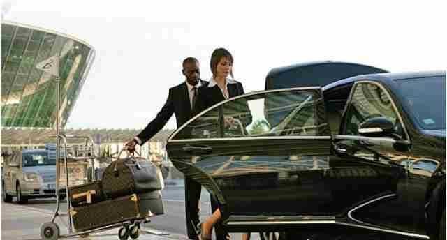 Two professionals in formal attire stand beside a black luxury sedan at an airport pickup area, with luggage and a trolley visible nearby