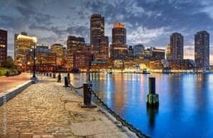 Boston skyline at dusk along the waterfront with illuminated high-rise buildings and a cobblestone promenade, reflecting in calm harbor waters