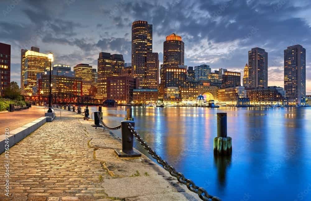 Boston skyline at dusk along the waterfront with illuminated high-rise buildings and a cobblestone promenade, reflecting in calm harbor waters
