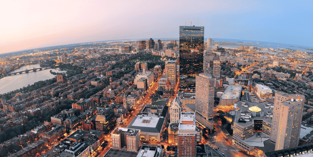Aerial view of Boston urban skyline at dusk, featuring a towering glass skyscraper, dense mid-rise blocks along the river, and warm orange-blue sunset lighting.