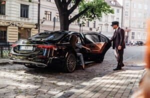 Lexington to Logan Airport Car Service with an open rear door and a professional chauffeur in a dark suit standing beside the car on a cobblestone street in Boston, near elegant townhouses and trees