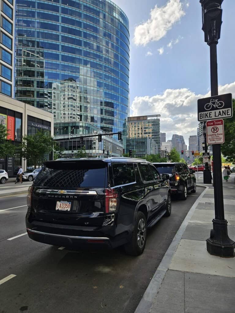 Black Suburban SUV on an urban Boston street beside modern glass buildings, reflecting sunlight, with a bike lane sign and city skyline