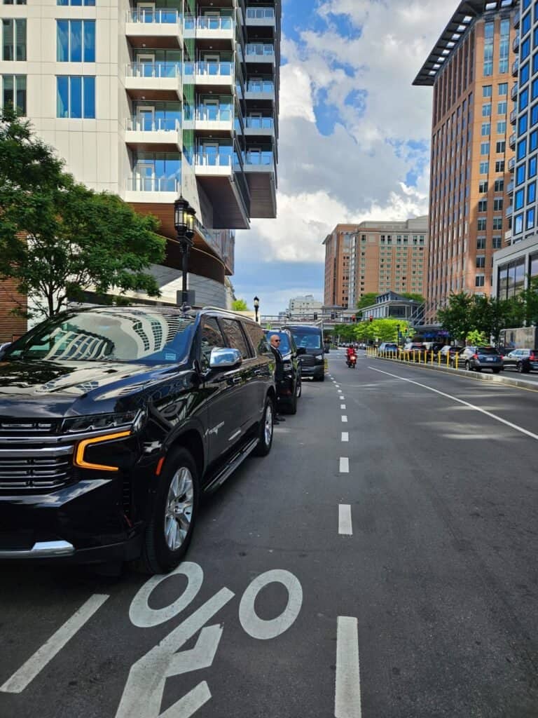 Boston Luxury Chauffeur black Chevrolet Suburbans lined up along a city street beside high-rise buildings, with a chauffeur standing by