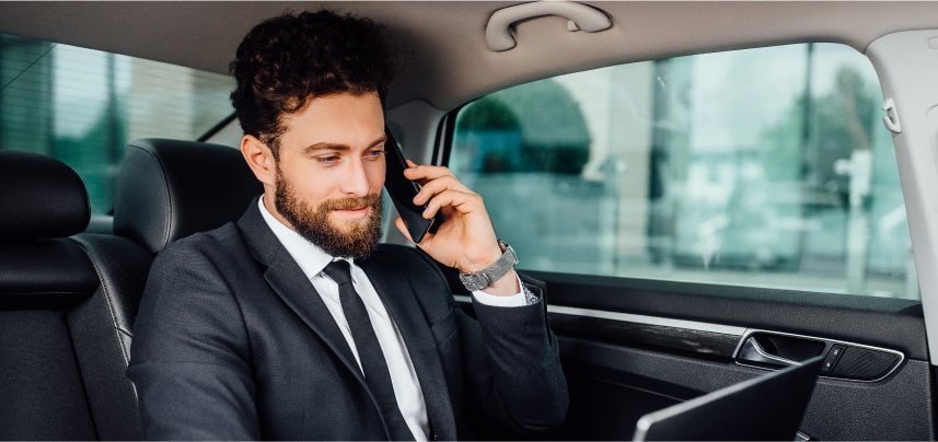 Boston Luxury Chauffeur wearing a tailored black suit sits in a premium sedan, calmly using a smartphone with a professional demeanor during a ride