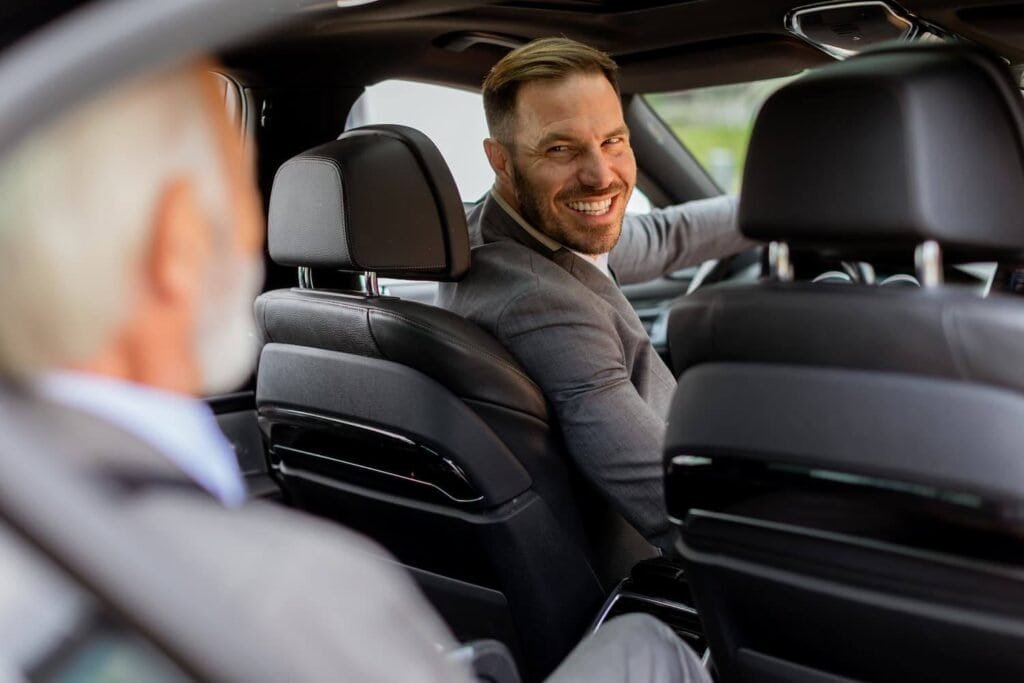 Chauffeur in a gray suit smiles from the back seat of a luxury sedan with black leather seats and a blurred driver in the foreground