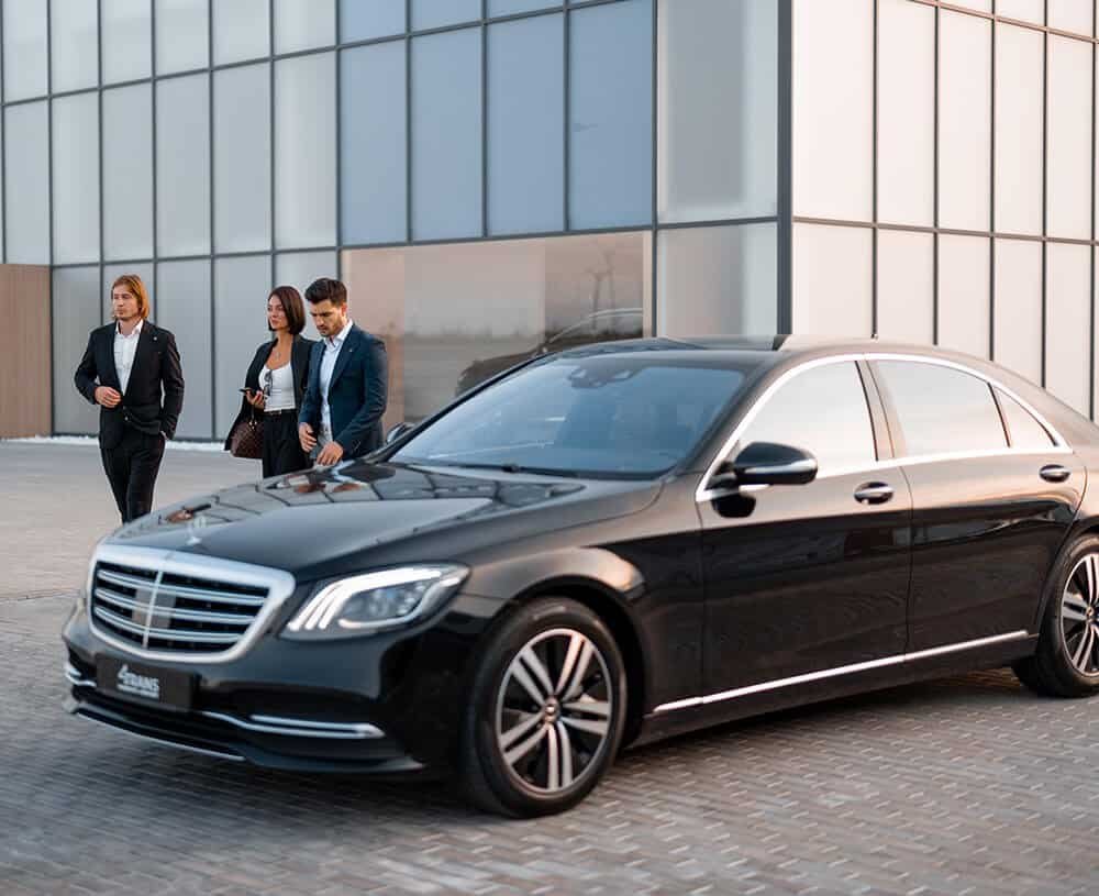 Boston Luxury Chauffeur black sedan parked outside a modern glass building while three executives in suits stand nearby, conveying premium corporate transportation in Boston
