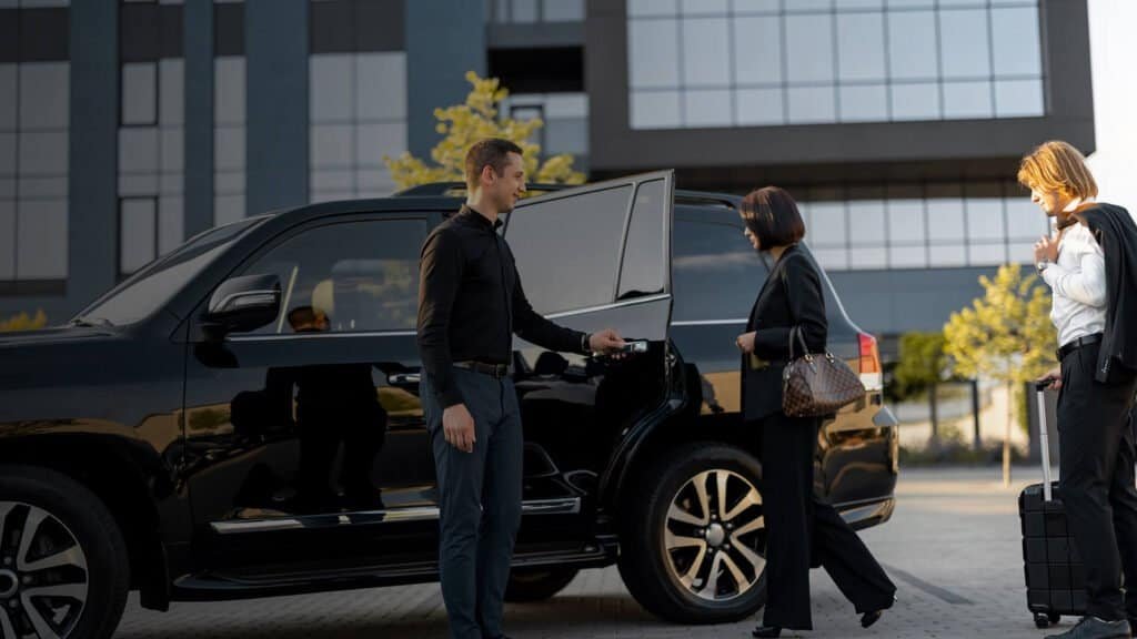 Black SUV with a smiling chauffeur opening the door for two guests in professional attire, alongside a white-capped traveler with a rolling case, in a modern business area