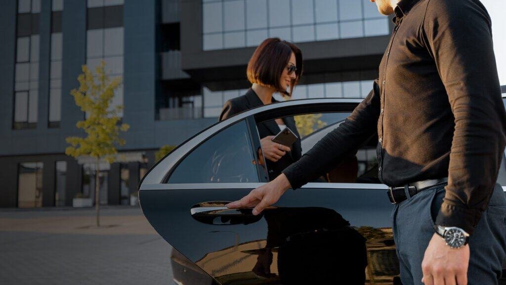 Black sedan with a chauffeur in a black shirt opening the door for a client in business attire; urban modern building backdrop.
