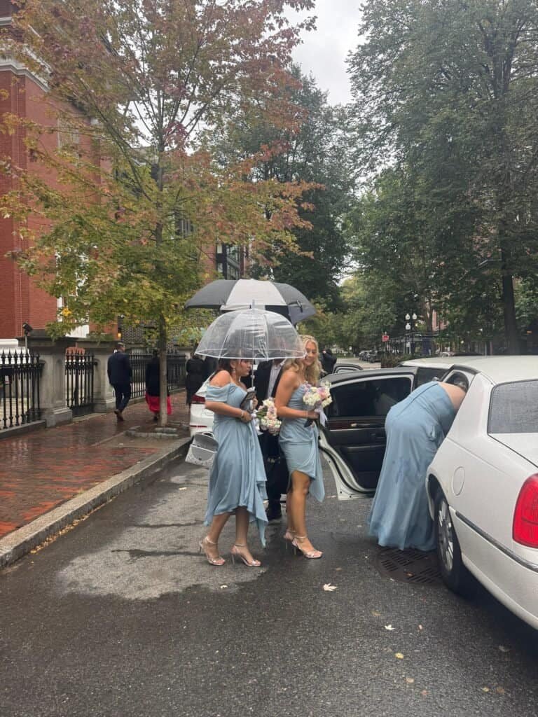 Group of women in light blue dresses carrying bouquets and clear umbrellas stepping from a white luxury sedan on a rainy city street with autumn trees