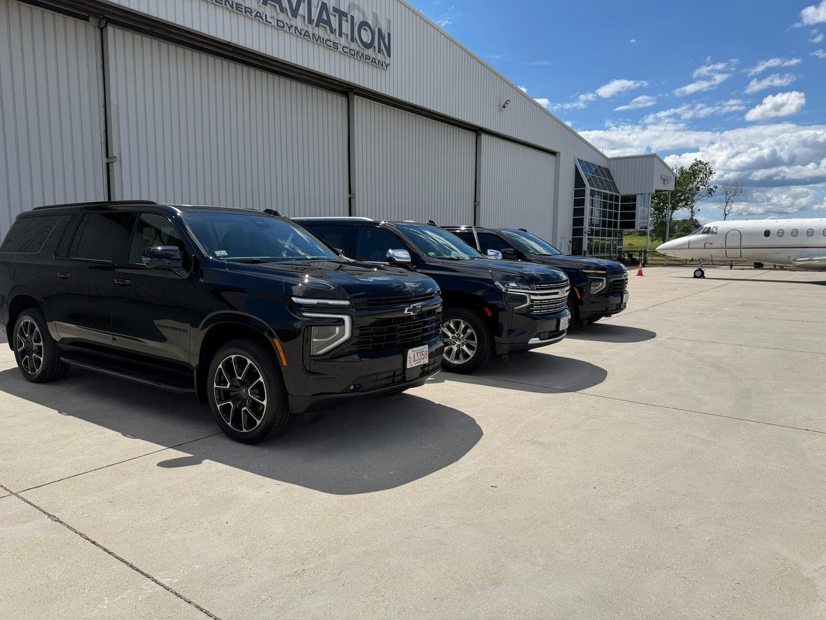 Boston Luxury Chauffeur fleet — three black Chevrolet Suburban SUVs lined up at Jet Aviation FBO at Hanscom Field with private jet in background